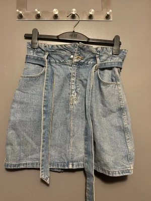 Blå jeanskjol med bälte från H&M - Snygg blå jeanskjol från H&M med tillhörande bälte i samma denimtyg. Kjolen har två stora fickor fram och stängs med knapp och dragkedja. Perfekt för dig som gillar en avslappnad men trendig stil.