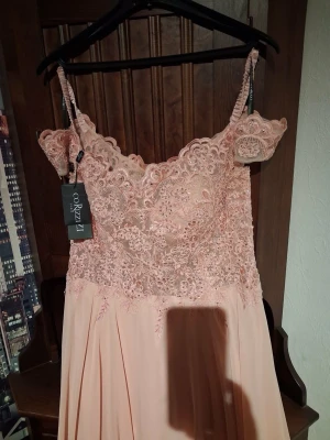 Lång ljusrosa spetsklänning Orizzzi - Elegant långklänning i ljusrosa från Orizzzi med vacker spets upptill och axelband. Klänningen har offshoulder-detaljer och ett svepande, flowy kjolparti. Perfekt för speciella tillfällen när du vill känna dig extra fin.
