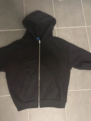 Svart zip hoodie med fickor - En klassisk svart hoodie med dragkedja framtill och två stora magfickor. Hoodien har en rymlig huva och ribbade muddar vid ärmslut och nederkant. Perfekt för en avslappnad stil och enkel att matcha med det mesta.