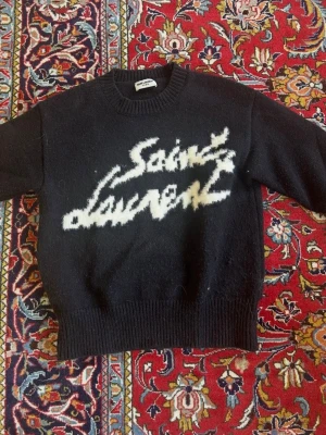 Svart Saint Laurent stickad tröja - Stickad svart tröja från Saint Laurent med vit fuzzy logga framtill i graffiti-stil. Tröjan har rund halsringning, ribbade muddar och nederkant. Materialet ser mjukt och lite luddigt ut, perfekt för en trendig och avslappnad look. Använd i en månad. 
