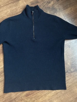 Mörkblå stickad halvzip tröja - Mörkblå stickad tröja med hög krage och dragkedja framtill. Tröjan har ribbad struktur och långa ärmar, perfekt för kyliga dagar. Enkel och stilren design som passar till det mesta.