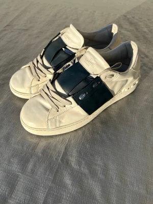 Valentino vita sneakers - Snygga vita sneakers från Valentino med bred marinblå läderdetalj över sidan och ovandelen. Klassisk låg modell med platta snören och diskreta nitar på hälen. Tillverkade i läder med gummisula. Perfekta för dig som gillar stilrena och exklusiva sneakers.