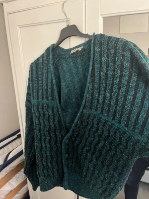 Grön stickad kofta från Tom Hanbury - Mysig stickad kofta i mörkgrönt och svart från Tom Hanbury. Koftan har ett vågigt mönster, djup V-ringning och knäppning framtill. Perfekt för lager på lager och ger en skön vibe till outfiten.
