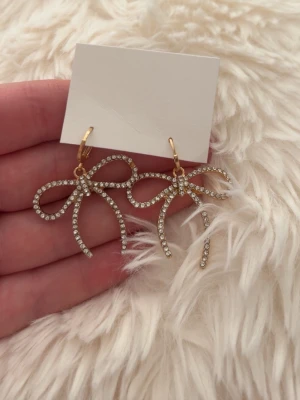 Guldiga rosettörhängen  -  örhängen i form av rosetter, dekorerade med gnistrande strass på framsidan och helt guldiga på baksidan. Örhängena är helt oanvända