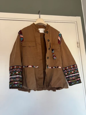 Brun jacka med broderade detaljer - Unik brun jacka med färgglada broderade partier på axlar och ärmar. Jackan har knappar framtill, två bröstfickor och en lätt utsvängd nederdel. 