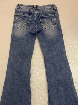 Blå bootcut jeans med slitningar - Säljer ett par blå bootcut jeans med snygga slitningar och markerade bakfickor med V-formade detaljer. Jeansen har låg midja, klassisk femficksdesign och är tillverkade i denim med lätt stretch. Perfekta för en avslappnad och trendig look.