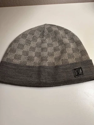 Grå Louis Vuitton mössa  - Snygg grå mössa från Louis Vuitton med klassiskt rutmönster och vikt kant. Diskret LV-logga på sidan ger en exklusiv touch. Tillverkad i mjukt stickat material som håller dig varm och stilren under kalla dagar.