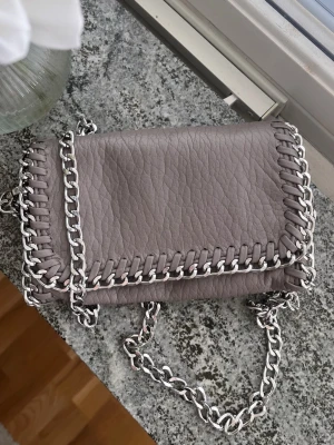 Handväska med kedjedetaljer - Grå snygg trendig handväska ifrån Scorett i modellen ”Tiamo pricilla S”. Väldigt lite använd och inget slitage på väskan, som ny. Kedjan går att ta av, göra kortare och ha lång.💘 Köpt för 500kr