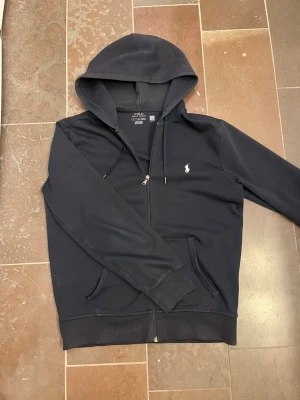 Mörkblå hoodie från Polo Ralph Lauren - Mörkblå hoodie med dragkedja från Polo Ralph Lauren. Klassisk design med vit broderad logga på bröstet, huva med dragsko och två fickor framtill. Perfekt för en chill och stilren look.