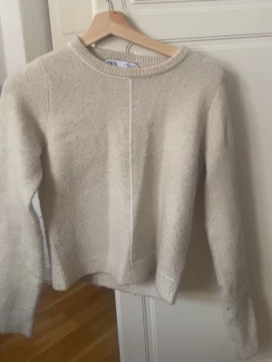 Beige stickad tröja från Zara - Mysig stickad tröja från Zara i beige färg med vit detalj på framsidan. Tröjan är långärmad och perfekt för att hålla sig varm under kyliga dagar💕