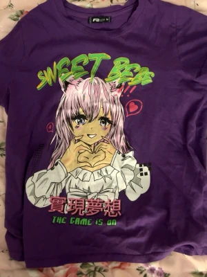 Lila anime t-shirt med tryck - Cool lila t-shirt från Fazspin med färgglatt anime-tryck på framsidan. Motivet visar en söt karaktär med kattöron och texten 'SWEET BEAST' och 'THE GAME IS ON' i neonfärger. Perfekt för dig som gillar japansk popkultur och unika prints. 