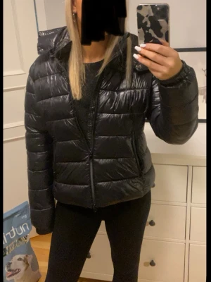 Svart pufferjacka från 157 - Snygg svart pufferjacka från 157 med glansig finish och quiltad design. Jackan har dragkedja framtill, hög krage och huva. Perfekt för kalla dagar när du vill hålla stilen och värmen. Passar dig som gillar en modern och enkel look.