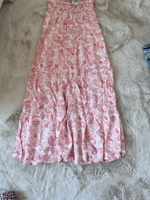 Långkjol med rosa paisleymönster - Säljer en flowy långkjol i vitt med rosa paisleymönster. Kjolen har resår i midjan och är gjord i ett lätt och luftigt material, perfekt för varma dagar. Passar dig som gillar boho-vibbar och vill ha något bekvämt men ändå snyggt. Passar super på mig som är ca 164,5! 