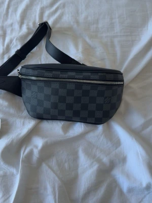 Axelväska Damier Graphite - Snygg axelväska i svart och grått Damier Graphite-mönster. Väskan har justerbar axelrem i läder, silverfärgad dragkedja och stilren, rektangulär form. Perfekt för dig som gillar exklusiva accessoarer och streetstyle.