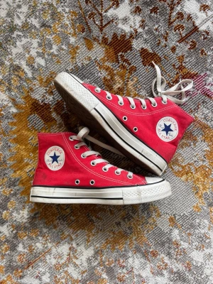 Röda Converse Chuck Taylor High - Klassiska röda Converse Chuck Taylor All Star high-tops med vit gummisula, vita snören och det ikoniska runda logotypmärket på sidan. Skorna är i canvas och har metallöljetter för snörningen. Perfekta för en cool och avslappnad stil.