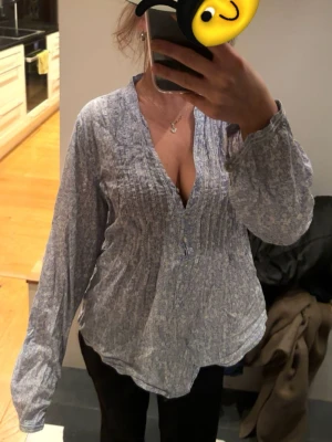 Ljusblå mönstrad blus med v-ringning - Ljusblå blus med diskret mönster och v-ringning. Blusen har knappar framtill och smockdetaljer över bröstet. Tillverkad i ett lätt och luftigt tyg som ger en avslappnad passform. Perfekt för dig som gillar en enkel men ändå stilren look.