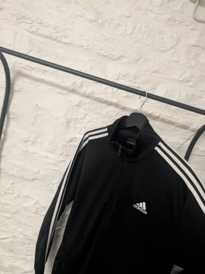 Adidas tröja - Snygg svart tröja från Adidas med dragkedja och klassiska vita ränder längs ärmarna. Adidas-logga på bröstet och hög krage. Tillverkad i mjukt och bekvämt material, perfekt för en sportig och avslappnad stil.