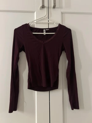 Vinröd ribbad långärmad topp - Snygg vinröd långärmad topp med v-ringning och spetsdetalj vid halsen. Ribbad struktur som ger en skön och stretchig passform. Perfekt att styla med jeans eller kjol för en trendig look.