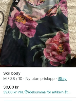 Blommig mesh body från iStay - Säljer en tunn och skir body från iStay med stort blommönster i rosa och grönt på mörkblå botten. Bodyn har meshmaterial som ger en lätt och luftig känsla, perfekt för att skapa en trendig lager-på-lager-look.