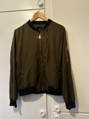 Olivgrön bomberjacka från Zara Basic - Snygg olivgrön bomberjacka från Zara Basic med svarta muddar och krage. Jackan har dragkedja framtill, fickor på sidorna och en ärmficka med dragkedja. Tillverkad i lätt, glansigt material som ger en cool streetkänsla.