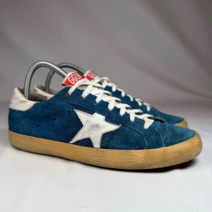  Golden Goose  - Snygga blå sneakers från Golden Goose med vit stjärna på sidan, vita skosnören och detaljer i vitt. Ovandelen är i mocka och sulan är beige/gulbrun. Röd etikett på plösen med GGDB-logga.