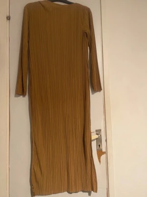 Plisserad långklänning - Säljer en långklänning med plisserat tyg och lång ärm. Klänningen har en rak passform och en slits på sidan för extra rörelsefrihet. Perfekt för dig som gillar stilrena och enkla plagg med en twist.