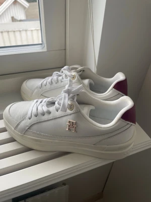 Vita sneakers från Tommy Hilfiger - Snygga vita sneakers från Tommy Hilfiger med guldfärgade detaljer. Skorna har klassisk låg profil, vita snören och är tillverkade i slätt lädermaterial. Perfekta för dig som vill ha en clean och stilren look.