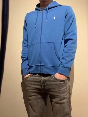 Blå zip hoodie från Ralph Lauren - Snygg blå hoodie med dragkedja från Ralph Lauren. Klassisk passform med huva och dragsko, två stora fickor framtill och den ikoniska vita loggan broderad på bröstet. Perfekt till jeans och t-shirt för en avslappnad look.