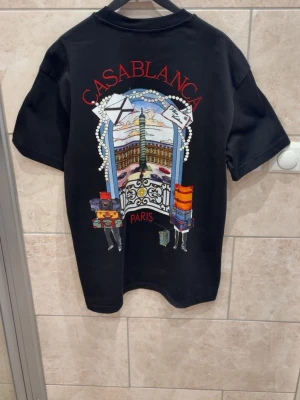 Svart Casablanca Paris t-shirt - Snygg svart t-shirt från Casablanca med färgglad grafisk print på ryggen som visar Paris och resväskor. T-shirten har rund hals och korta ärmar. Perfekt för dig som gillar unika prints och streetwear-vibe. Helt oanvänd