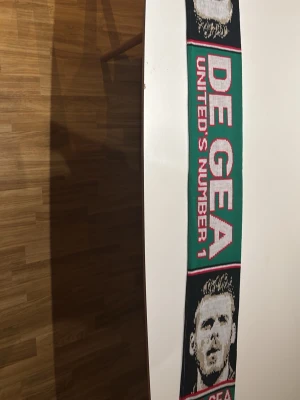 Grön De Gea halsduk United - Säljer en grön halsduk med texten 'DE GEA UNITED'S NUMBER 1' i vitt och rött. Halsduken har porträttmotiv och svarta detaljer. Perfekt för dig som är fotbollsfan och vill visa ditt stöd för De Gea och United.