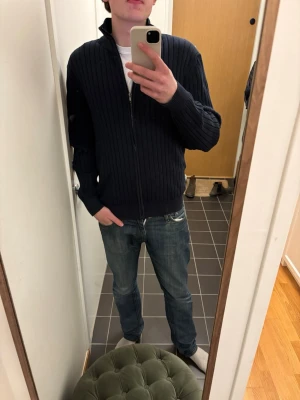 Mörkblå ribbad ziptröja - Säljer en mörkblå ribbstickad tröja med dragkedja framtill och hög krage. Tröjan har långa ärmar och en clean, stilren look som passar perfekt till jeans. Materialet ser ut att vara mjukt och stretchigt, vilket gör den bekväm att bära.