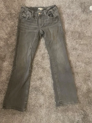 Grå bootcut jeans - Säljer ett par grå bootcut jeans med snygga fickdetaljer och knappar bak. De är knappt använda och har inge defekter! De är storlek 170 från Gina!