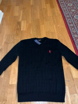 Svart kabelstickad tröja Polo Ralph Lauren - Kabelstickad svart tröja från Polo Ralph Lauren med rund halsringning och röd broderad logga på bröstet. Tröjan har ribbade muddar vid ärmslut och nederkant. Klassisk och stilren design som passar till mycket.