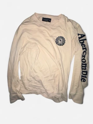 Beige långärmad tröja Abercrombie - Långärmad beige tröja från Abercrombie & Fitch med tryckt logga på bröstet och stor text längs ena ärmen. Tillverkad i mjuk bomull och har en avslappnad passform. Perfekt för en casual och trendig look.