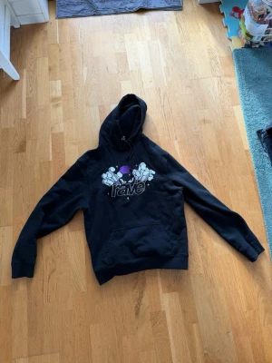 Svart hoodie från Rave Skateboards - Svart hoodie från Rave Skateboards med tryck framtill i vitt och lila. Klassisk känguruficka och justerbar huva med dragsko. Skön och avslappnad passform, perfekt för skate eller chill. Materialet är mjukt och känns bekvämt mot huden.