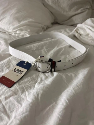 Vitt skärp från Tommy Jeans - Snyggt vitt skärp från Tommy Jeans med präglad logga längs bandet. Skärpet har ett klassiskt silverfärgat spänne och detaljer i rött och blått vid spännet. Perfekt accessoar för att lyfta din outfit.