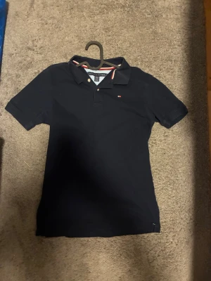 Mörkblå Tommy Hilfiger Piké  - Klassisk Mörkblå pikétröja från Tommy Hilfiger med korta ärmar och liten logga på bröstet. Kragen har en snygg vit och röd kant som ger en sportig touch. Liknande moncler pikén som är stilren och perfekt för sommarn.