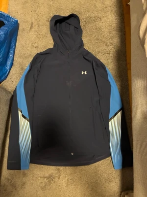 Under Armour Windbreaker  - Snygg vindjacka från Under Armour i mörkblått med ljusblå och vita detaljer på ärmarna. Jackan har hel dragkedja, huva och långa ärmar med sportig design. Perfekt för aktiva dagar och har logotypen på bröstet. Använd 2 gånger! Vid behov av fler frågor eller byten hör av er!