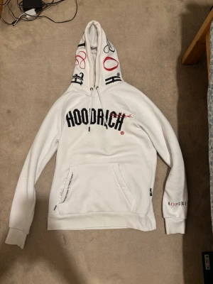 Vit hoodie från Hoodrich med tryck - Säljer en vit hoodie från Hoodrich med svart och detaljer på huvan. Hoodien har en stor ficka . Den är väldigt skön och ser ut som helt ny.