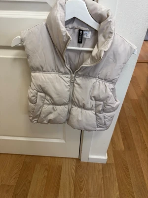 Beige dunväst från Hm - Snygg beige dunväst från Hm med hög krage och dragkedja framtill. Västen har två stora fickor på sidorna och en puffig, quiltad design som ger en trendig look. Perfekt att slänga över en hoodie eller tröja för extra värme. Stl xs 