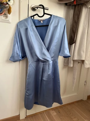 Ljusblå omlottklänning i satin - Snygg ljusblå omlottklänning i glansigt satinmaterial med v-ringning och halvlånga ärmar. Klänningen har ett band i midjan för att skapa en fin siluett och en elegant look. Använd 1 gång 