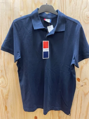 Marinblå pikétröja från Clique - Snygg marinblå pikétröja från Clique med klassisk krage och korta ärmar. Tillverkad i bomull för en skön känsla och stilren look. Perfekt till jeans eller shorts för en enkel och clean outfit.