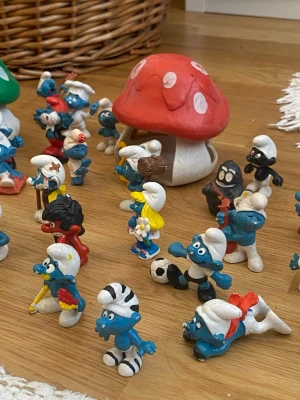 Samling Smurf-figurer och svamphus - Säljer en samling Smurf-figurer i plast med olika utstyrslar och tillbehör, samt två färgglada svamphus i rött och grönt. Figurerna är blå med vita, röda och svarta detaljer, vissa har accessoarer som fotboll, blommor och verktyg. Perfekt för dig som gillar retro och samlarprylar.