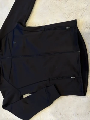 Svart träningsjacka från Craft - Svart träningsjacka från Craft med hel dragkedja framtill och två sidofickor med dragkedja. Tillverkad i ett stretchigt funktionsmaterial som passar perfekt för löpning eller gym. Diskret logga på bröstet och smal passform.