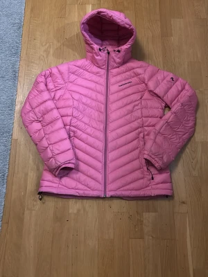 Rosa dunjacka Peak Performance dam XL - Snygg rosa dunjacka från Peak Performance i storlek XL. Jackan har huva, dragkedja framtill och två sidofickor med dragkedja. Quiltad design med diagonala sömmar ger en modern look. Perfekt för kalla vinterdagar och riktigt varm.