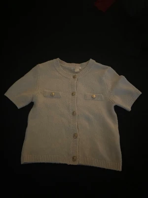 Vit kortärmad kofta från Gina Tricot - Kortärmad vit kofta från Gina Tricot med rund halsringning och dekorativa guldknappar framtill. Två fejkfickor med knappdetaljer på bröstet och ribbade kanter. Perfekt för lager på lager och enkel att matcha. Den har inte varit andvänd mer än 5 gånger💕 tröjan kostade nått med 200 men kan sänka priset 💓