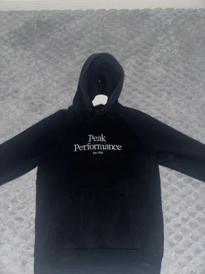 Svart hoodie från Peak Performance - Snygg svart hoodie från Peak Performance med vit broderad logga och texten 'Est. 1986' på bröstet. Klassisk känguruficka framtill och justerbar huva utan snören de är bort tappade.