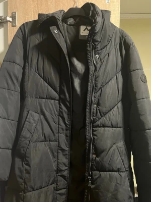 Svart lång dunjacka från Virtus - Snygg svart lång dunjacka från Virtus med hög krage och dragkedja framtill. Jackan har diskreta sidofickor och ett stilrent märke på ärmen. Perfekt för kalla vinterdagar tack vare det vadderade materialet och den klassiska designen.