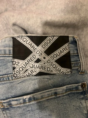 Ljusblå Dsquared2 jeans med patch - Säljer ett par ljusblå jeans från Dsquared2 med en stor svart och vit patch bak i midjan med logga. Jeansen har raka ben, klassisk femficksmodell och coola färgstänk för en trendig look. Perfekta för dig som gillar streetwear och vill sticka ut.