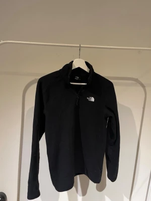Svart fleece från The North Face - Mysig svart fleece från The North Face med halv dragkedja och hög krage. Perfekt för kalla dagar och enkel att slänga på sig. Diskret logga broderad på bröstet. Tillverkad i mjukt fleecematerial och har långa ärmar.